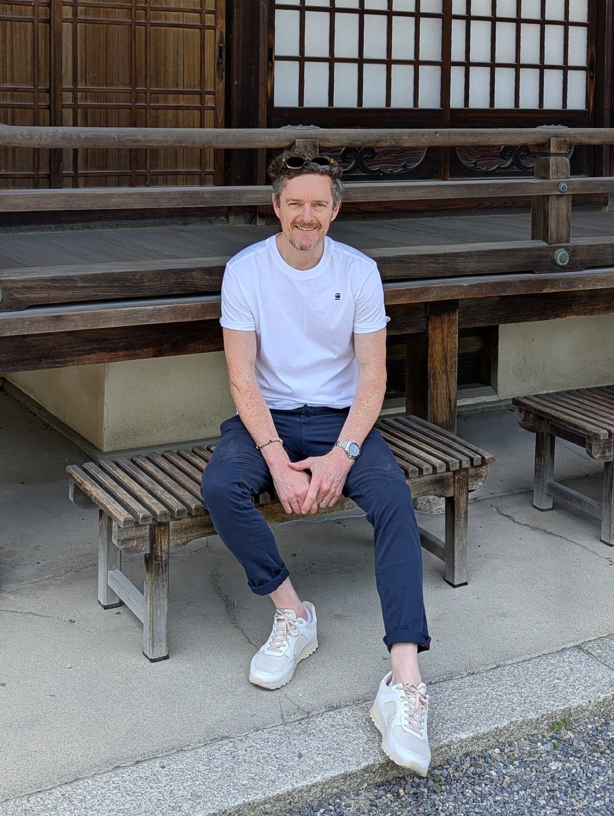 Tom Vining sitting on a wooden bench outside a temple in Japan, in a white T-shirt and navy trousers